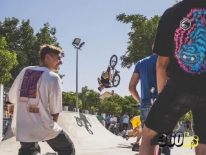 Grupo de riders en park con sus bicis BMX , de fondo aparece un rider haciendo un truco en el aire.