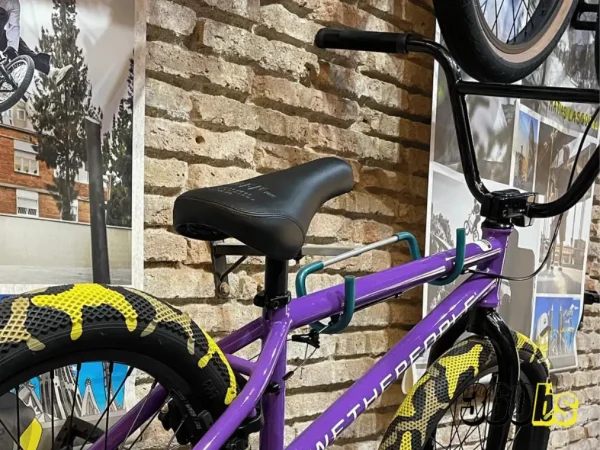 Bicicleta BMX colorida con cuadro morado, llantas negras y sillín BMX cómodo, expuesta frente a una pared con fotos de acción de ciclistas BMX al fondo.