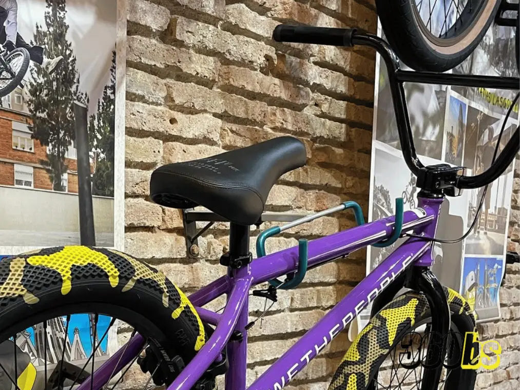 Bicicleta BMX colorida con cuadro morado, llantas negras y sillín BMX cómodo, expuesta frente a una pared con fotos de acción de ciclistas BMX al fondo.