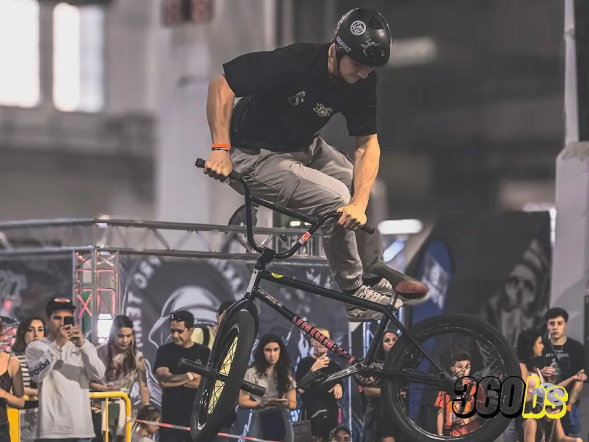 Ciclista realizando trucos BMX en un skatepark indoor ante una multitud.