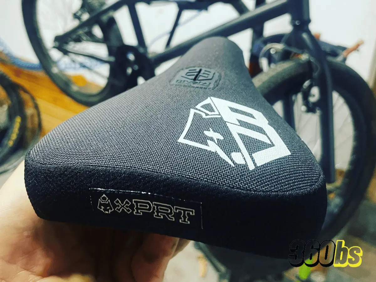 Primer plano de un sillín de BMX negro con el logotipo de Expert y emblemas blancos, sostenido por una mano, con una bicicleta BMX desenfocada al fondo. 